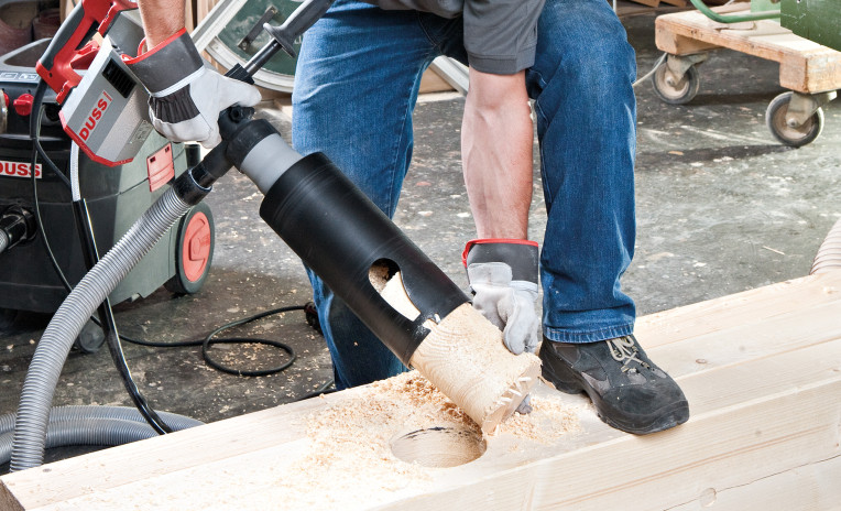 Professional woodworking machine drilling a large hole in a timber beam using a wood cutter tool, producing wood chips during structural timber processing